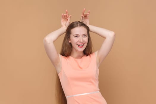 Portrait of funny smiling joyful woman with long hair standing with raised arms, showing horns, looking at camera, wearing elegant dress. Indoor studio shot isolated on brown background.