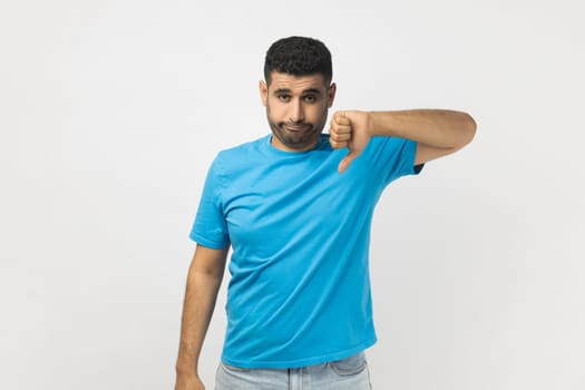 Portrait of unshaven man wearing blue T- shirt standing assess project, shows sign of dislike, looks with negative expression and disapproval. Indoor studio shot isolated on gray background.