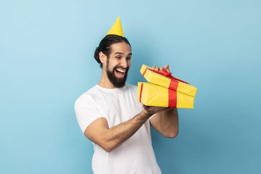 Portrait of extremely happy man wearing white T-shirt and party cone looking into gift box, opening present and peeking inside with happiness. Indoor studio shot isolated on blue background.