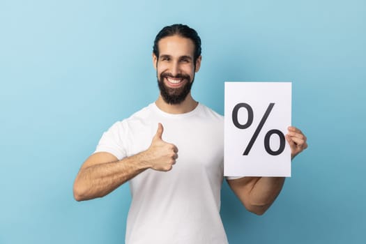 Portrait of positive smiling man with beard wearing white T-shirt holding and pointing paper with percent sign inscription, showing thumb up. Indoor studio shot isolated on blue background.