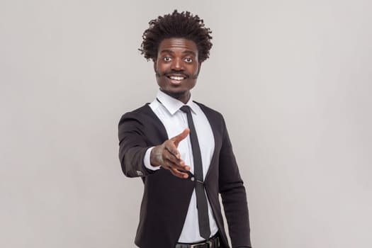 Friendly man with petit goatee, giving hand for handshake as sign of greeting, smiling widely and sincerely, looking very glad to see person he meeting. Indoor studio shot isolated on gray background.