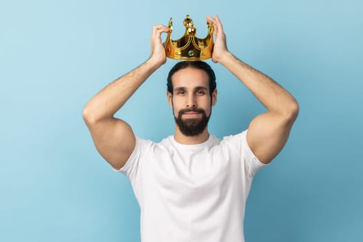 Portrait of serious independent man with beard in white T-shirt wearing golden crown, looking with arrogance and confidence, privileged status. Indoor studio shot isolated on blue background.