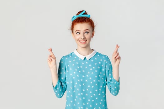 Portrait of smiling attractive hopeful ginger woman with bun hairstyle, standing with crossed fingers, making wish, wearing blue dress. Indoor studio shot isolated on gray background.