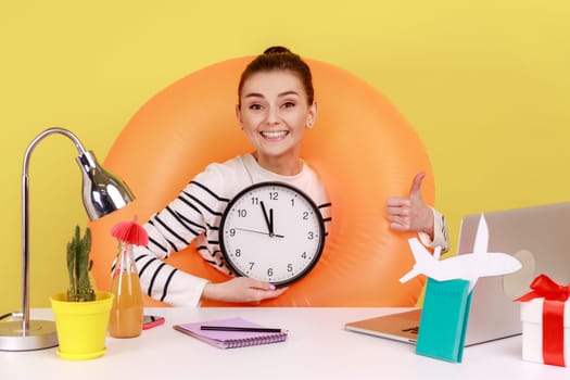 Happy excited woman in rubber ring on neck sitting at workplace with laptop, showing wall clock, time to go on summer vacation. Indoor studio studio shot isolated on yellow background.