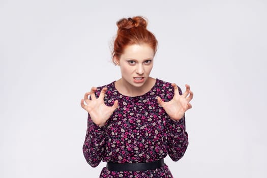 Portrait of angry aggressive young adult ginger woman wearing dress standing with raised arms and clenching teeth, expressing hate and anger. Indoor studio shot isolated on gray background.