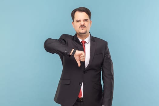 Displeased sad showing man with mustache standing showing dislike gesture, grimacing, feels dissatisfied, wearing black suit with red tie. Indoor studio shot isolated on light blue background.