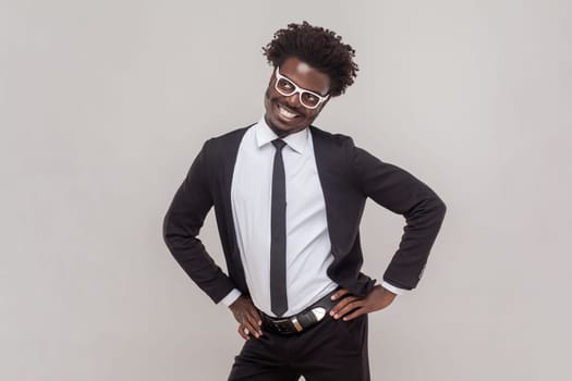 Portrait of funny positive man in glasses standing with hands on hips, looking at camera with toothy smile, wearing white shirt and tuxedo. Indoor studio shot isolated on gray background.