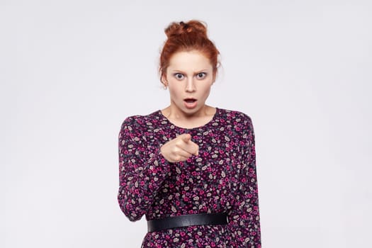 Portrait of extremely astonished ginger woman wearing dress pointing at camera, being very shocked to choose you, keeps mouth open. Indoor studio shot isolated on gray background.