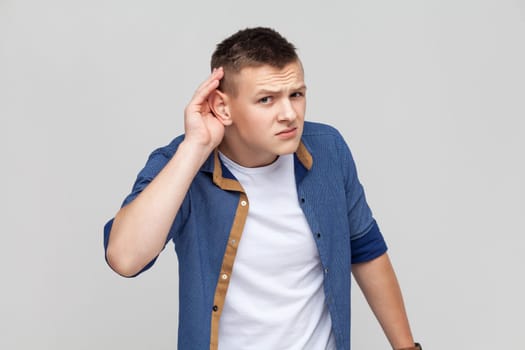 Portrait of teenager boy wearing blue shirt holding hand near ear and listening carefully, having hearing problems, deafness in communication. Indoor studio shot isolated on gray background.