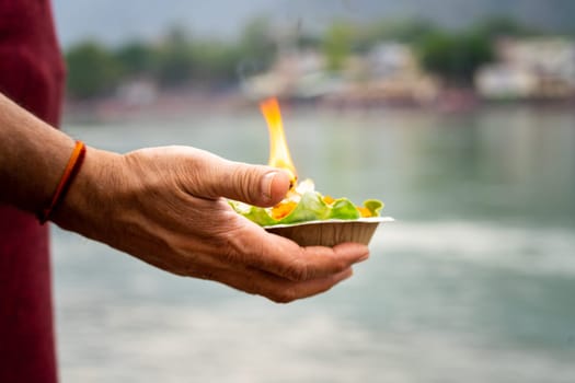 shallow depth of feild shot of man holding leaf plate cup with marigold gande flowers rose and a lit oil ghee lamp diya as an offering to the river ganga at rishikesh