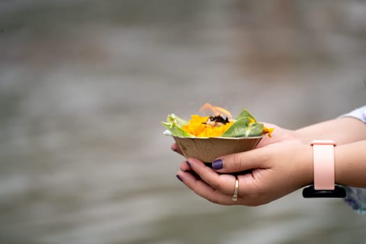 shallow depth of feild shot of woman with watch holding leaf plate cup with marigold gande flowers rose and a lit oil ghee lamp diya as an offering to the river ganga at rishikesh India