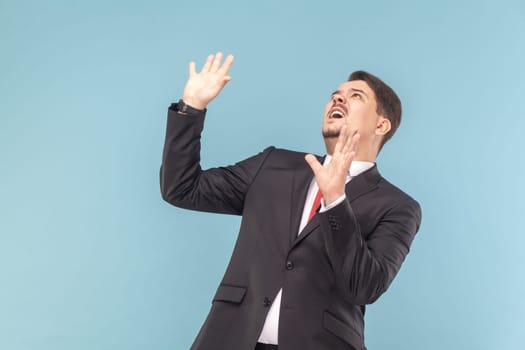 Portrait of sacred frighten man with mustache standing looking up, showing stop gesture, feels danger, wearing black suit with red tie. Indoor studio shot isolated on light blue background.