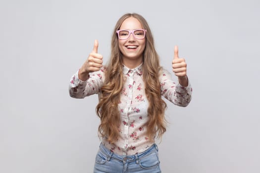Woman in glasses with wavy blond hair looking at camera with toothy smile and showing thumbs up, approval sign, satisfied with service, good feedback. Indoor studio shot isolated on gray background.