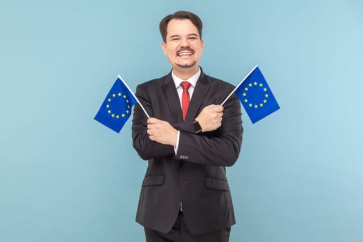 Portrait of man holding European flags, symbol both the European Union and the identity and unity of Europe, wearing black suit with red tie. Indoor studio shot isolated on light blue background.
