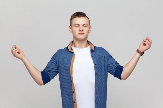 Portrait of calm meditative handsome teenager boy wearing blue shirt standing with closed eyes and yoga gesture pose, trying to calm down. Indoor studio shot isolated on gray background.