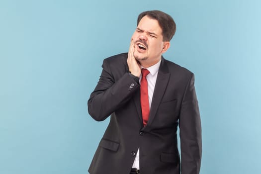 Portrait of man suffers painful toothache, has rotten tooth, needs to visit dentist, keeps eyes closed from pain, wearing black suit with red tie. Indoor studio shot isolated on light blue background.