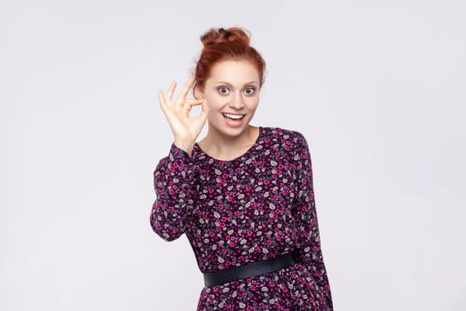 Portrait of delighted joyful cheerful young adult redhead woman wearing dress showing ok sign, looking at camera with toothy smile. Indoor studio shot isolated on gray background.