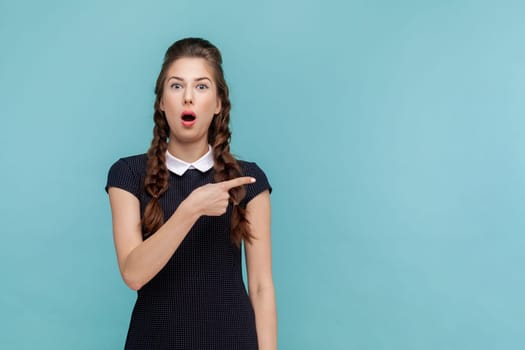 Portrait of shocked surprised woman points away on copy space, suggests follow this direction or click on link, wearing black dress. woman Indoor studio shot isolated on blue background.