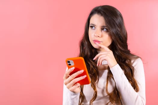 Portrait of thoughtful attractive young brunette woman with a smartphone, chatting with friends looking up empty space or thinking about new post isolated on pink color background.
