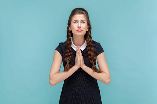 Portrait of hopeful uneasy woman with braids begging for forgiveness keeps palms in praying gesture, asks mercy and says please, needs your help. woman Indoor studio shot isolated on blue background