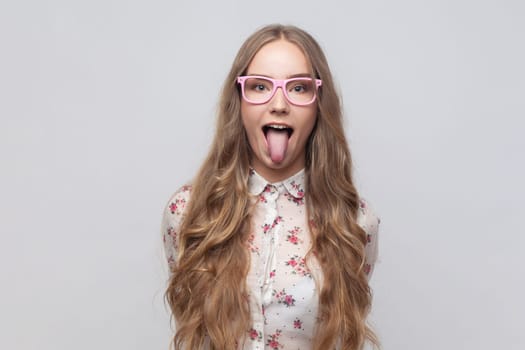 Portrait of childish carefree woman in glasses with long blond hair showing out tongue and closing eyes with naughty disobedient grimace, making face. Indoor studio shot isolated on gray background.