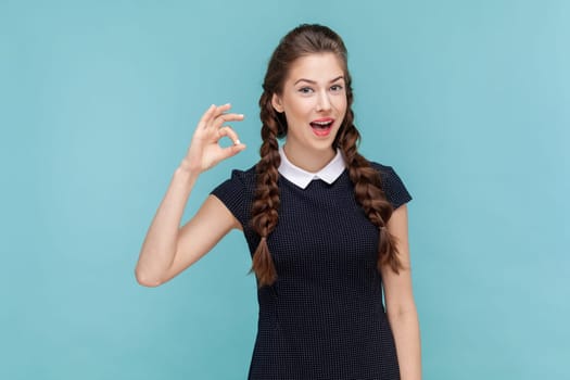 Portrait of beautiful woman with braids making okay gesture, demonstrates agreement, likes idea, smiles happily, wearing black dress. woman Indoor studio shot isolated on blue background.