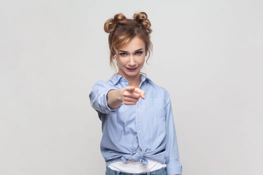 Portrait of joyous pretty blonde woman pointing finger to camera, expresses choice, smiling broadly, saying you are chosen, wearing blue shirt. Indoor studio shot isolated on gray background.