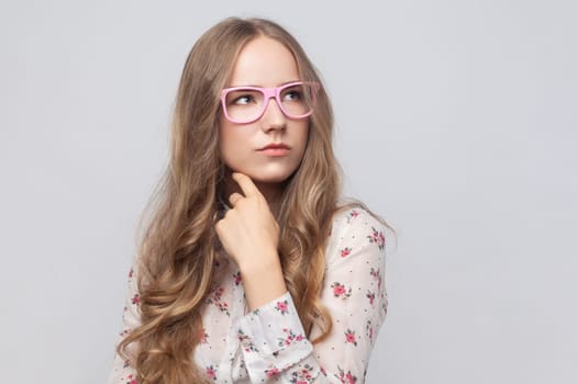 Portrait of thoughtful pensive woman in glasses with long blond hair thinking about future, holding chin, having serious facial expression. Indoor studio shot isolated on gray background.