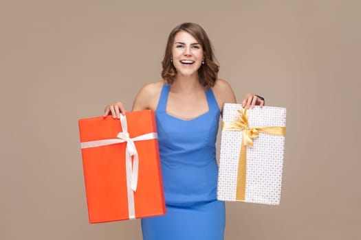 Portrait of extremely happy positive woman with wavy hair standing with two shopping bags, being glad to do shopping, wearing blue dress. Indoor studio shot isolated on light brown background.