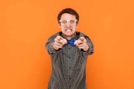 Portrait of angry aggressive carazy man nerd pointing at you, looking at camera with clenched teeth, wearing shirt with blue bow tie and white glasses. Indoor studio shot isolated on orange background