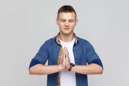 Portrait of calm attractive smiling teenager boy wearing blue shirt standing in yoga pose and try to relaxing, keeps palms together. Indoor studio shot isolated on gray background.