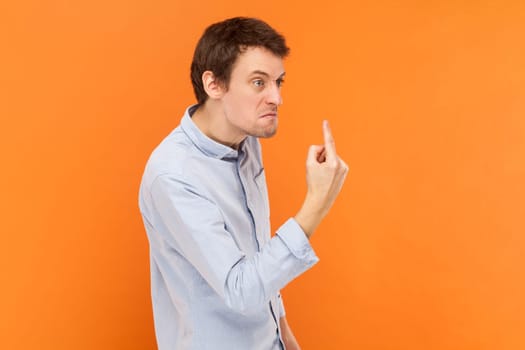 Side view portrait of angry rude man standing looking away with aggression, showing middle finger, fighting with someone, wearing light blue shirt. Indoor studio shot isolated on orange background.