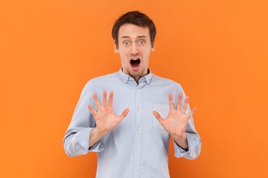Portrait of scared frighten young adult man standing with raised arms, showing stop gesture, sees something scary, wearing light blue shirt. Indoor studio shot isolated on orange background.