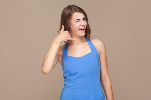 Call me back sign. Portrait of woman with wavy hair makes telephone gesture, asks to call her, expresses positive emotions, wearing blue dress. Indoor studio shot isolated on light brown background.