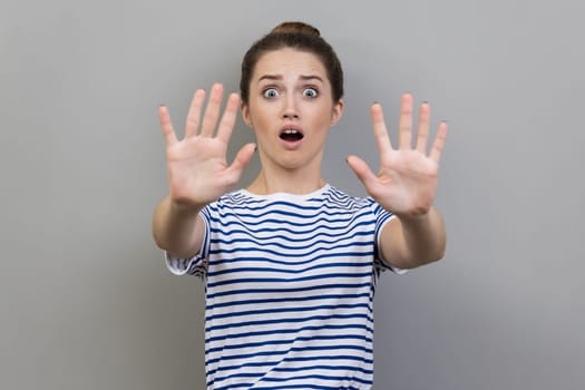 Portrait of scared young adult woman wearing striped T-shirt gesturing stop with palms and looking surprised with frightened eyes. Indoor studio shot isolated on gray background.