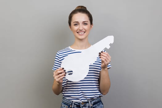 Portrait of happy woman wearing striped T-shirt holding big white house key, real estate advertisement, house purchase, apartment rent. Indoor studio shot isolated on gray background.