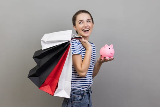 Portrait of excited positive woman wearing striped T-shirt holding shopping bags and piggy bank, cashback from buying purchases. Indoor studio shot isolated on gray background.
