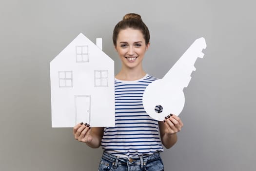 Portrait of satisfied pretty positive woman wearing striped T-shirt holding paper house and big key, real estate purchase, rental services, mortgage. Indoor studio shot isolated on gray background.