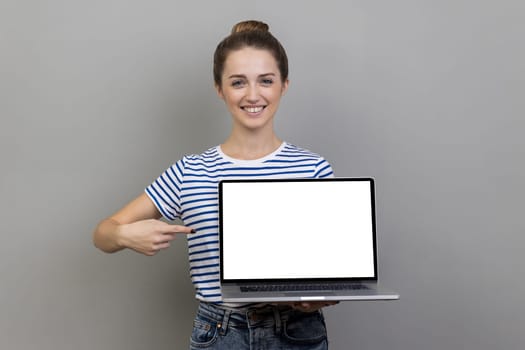 Portrait of woman wearing striped T-shirt pointing finger at laptop with white blank display, showing empty screen, bragging with internet advertisement. Indoor studio shot isolated on gray background