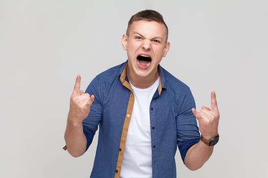 Yeah, awesome. Portrait of teenager boy wearing blue shirt showing rock and roll hand gesture, crazy devil horns with fingers and yelling. Indoor studio shot isolated on gray background.