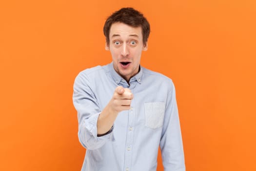 Portrait of shocked surprised man standing and looking at camera with amazed expression, pointing at you, choosing you, wearing light blue shirt. Indoor studio shot isolated on orange background.