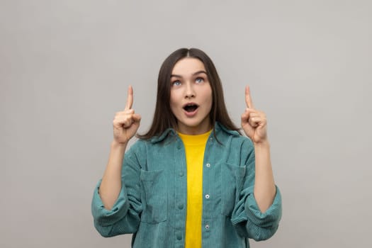 Portrait of surprised woman with open mouth pointing fingers up at empty copy space, place for advertisement, wearing casual style jacket. Indoor studio shot isolated on gray background.