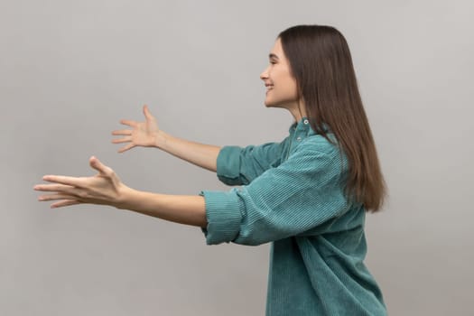 Come into my arms, free hugs. Side view of adorable hospitable woman smiling and reaching out hands, going to embrace, wearing casual style jacket. Indoor studio shot isolated on gray background.