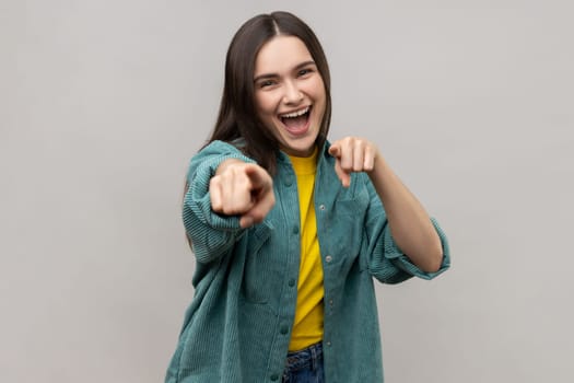 Hey you. Positive dark haired young woman pointing to camera, showing direction and choosing, indicating with index finger, wearing casual style jacket. Indoor studio shot isolated on gray background.