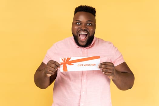 Portrait of excited amazed happy man wearing pink shirt holding gift voucher, looking at camera with positive expression, winning present. Indoor studio shot isolated on yellow background.