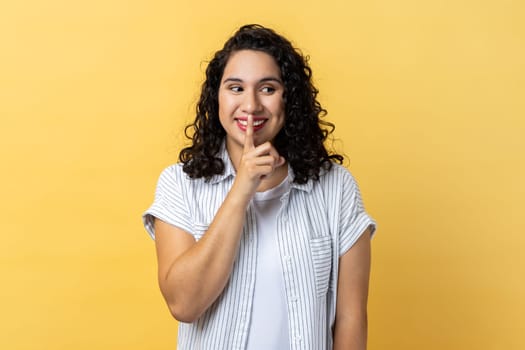 Portrait of attractive woman with dark wavy hair standing, showing hush sign and looking away with toothy smile, sharing secret. Indoor studio shot isolated on yellow background.