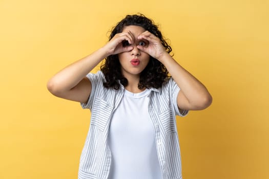 Portrait of woman with dark wavy hair spying, watching unbelievable shocking event, looking through fingers imitating binoculars. Indoor studio shot isolated on yellow background.