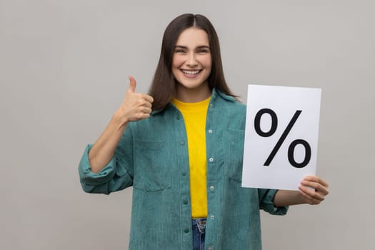 Smiling woman holding and showing paper percent sign inscription, looking at camera and showing thumb up, wearing casual style jacket. Indoor studio shot isolated on gray background.