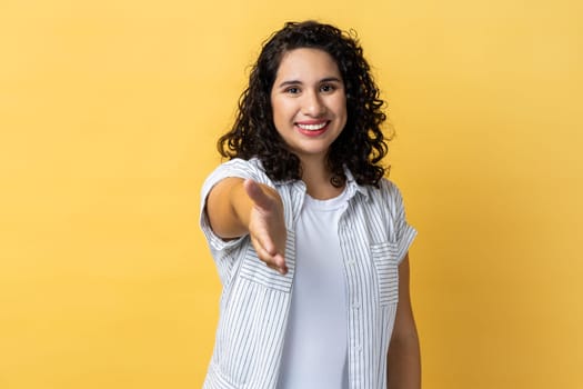 Let me introduce myself. Portrait of woman with dark wavy hair friendly woman giving hand to handshake, greeting guests with toothy smile. Indoor studio shot isolated on yellow background.