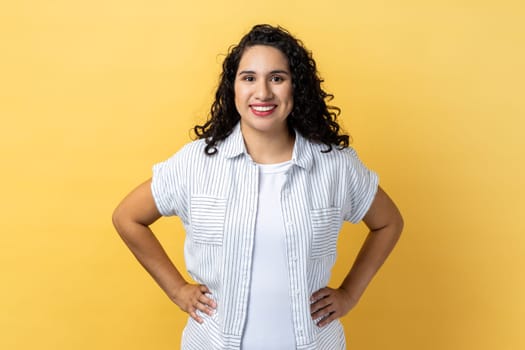 Portrait of friendly positive woman with dark wavy hair standing with hands on hips, looking at camera with satisfied facial expression. Indoor studio shot isolated on yellow background.